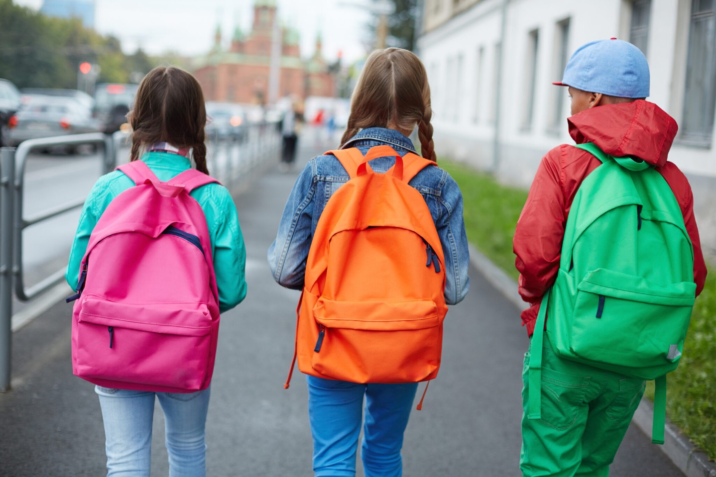Three children walking down the street, each wearing a brightly colored backpack.